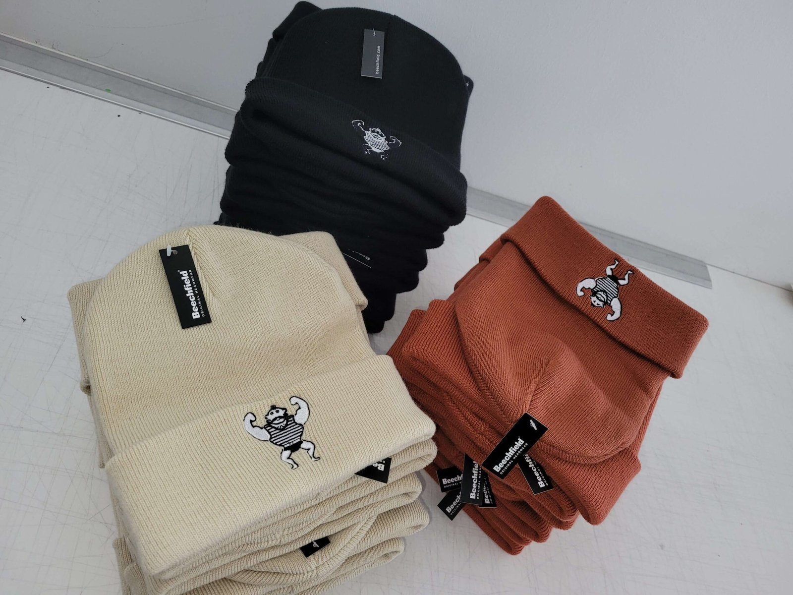 Gestapelte Beanies in Schwarz, Naturweiß und Terrakotta mit gesticktem Kundenlogo auf dem Werkstatttisch
